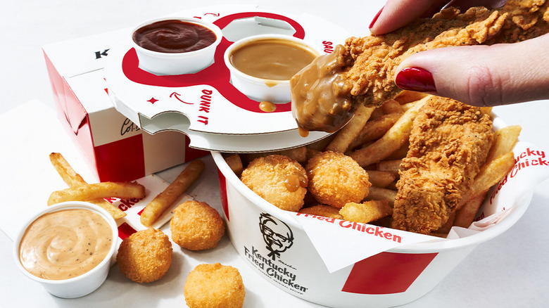 A woman with red nail polish lifts a KFC chicken tender after dipping it in sauce