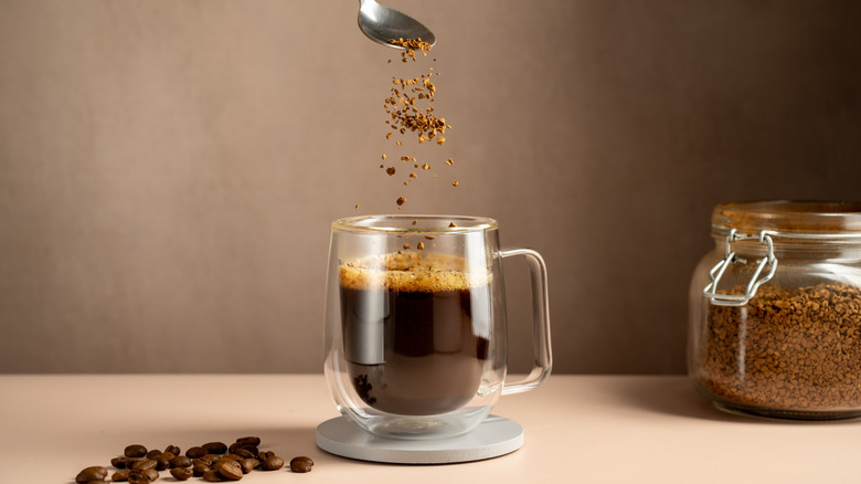 Instant coffee granules fall from a spoon into a cup of coffee, which rests beside some coffee beans.