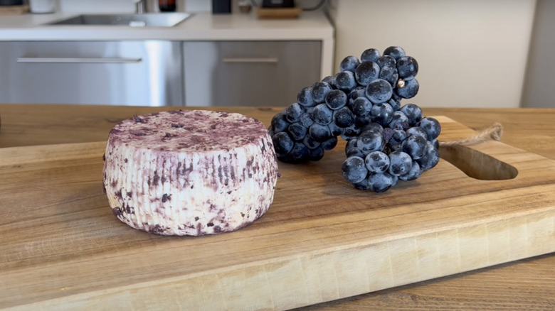 Wheel of drunk cheese next to wine grapes on cutting board