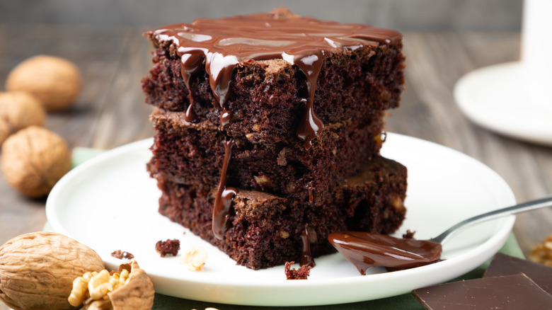Three brownies stacked on a plate with chocolate sauce on top and scattered whole walnuts