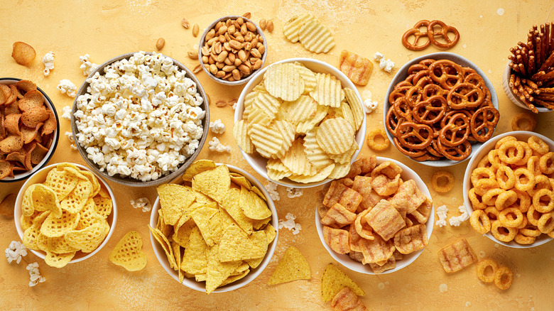 Assorted chips, pretzels, and popcorn in bowls on orange surface