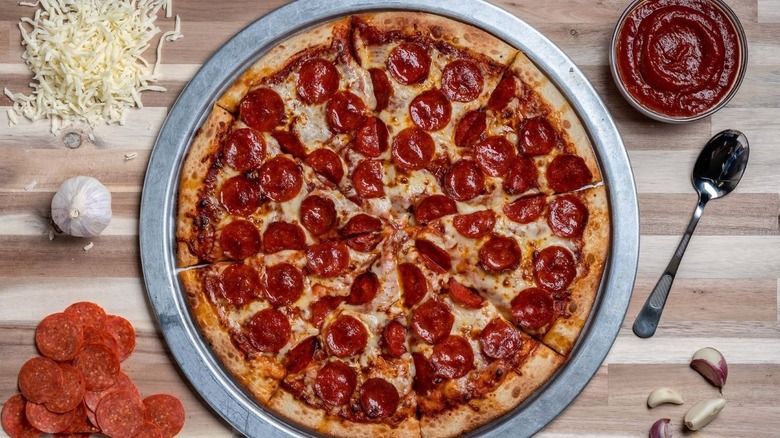 Top view of pepperoni pizza surrounded by cup of sauce, shredded cheese, garlic, and pepperoni slices