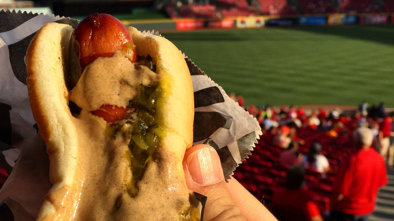 A hand holds a hot dog covered with brown mustard, with a ball stadium in the background