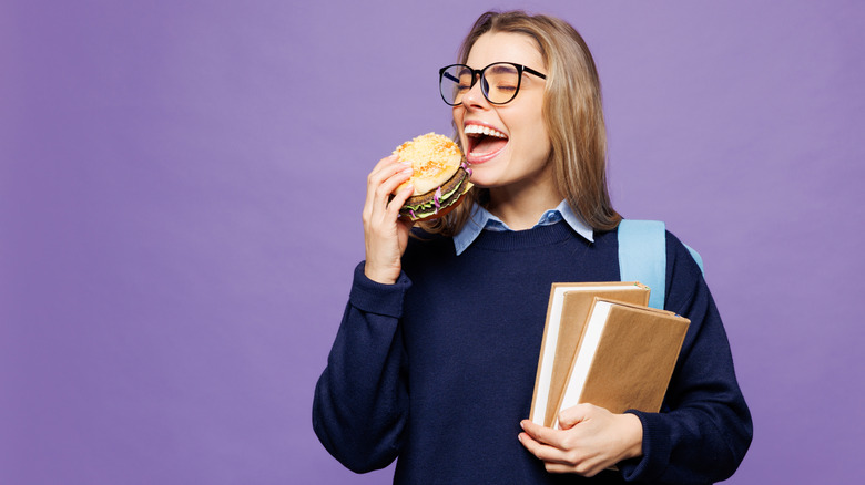 An adult student wearing glasses and holding books bites into a hamburger.