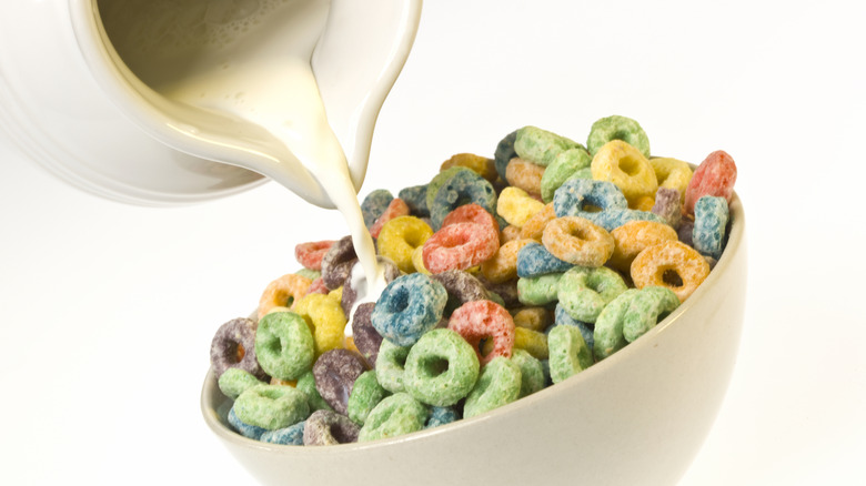 Milk is poured from a white jug onto a bowl of colorful cereal