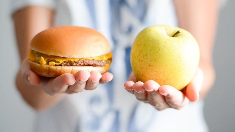 Hands holding one cheeseburger and one apple