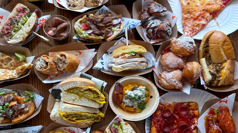 An assortment of different foods rests on a table, all from Citi Field, including pizza, chicken wings, sandwiches, hamburgers, donuts, wraps, steak, ad more