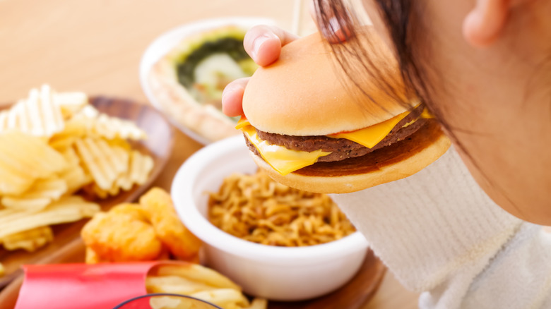 A diner holds up a cheeseburger to take a bite, with other processed foods like Asian noodles, french fries, chicken nuggets, and potato chips sitting on the table