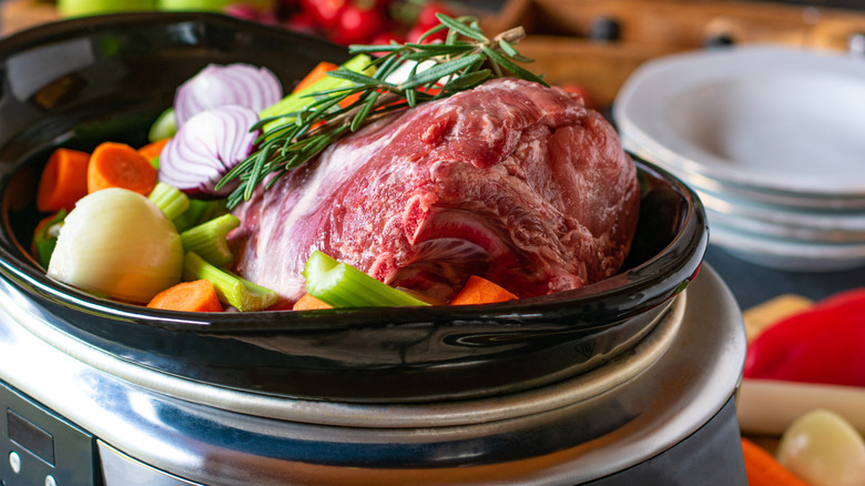 A large slab of meat sits in a slow cooker with onions, rosemary springs, carrots, and celery