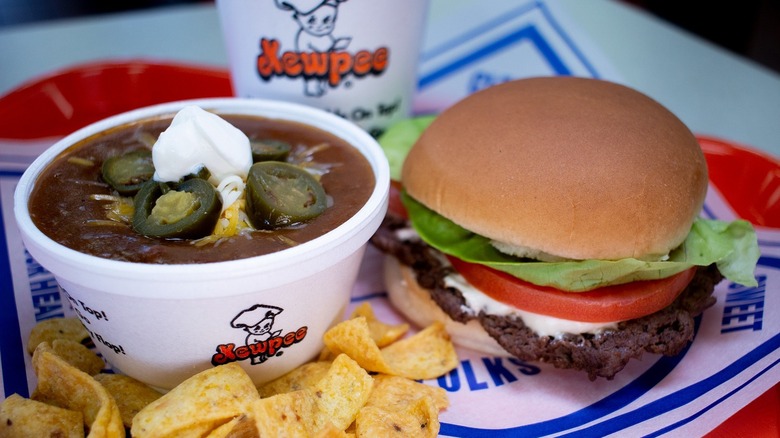 A Kewpee hamburger sits on a restaurant tray with a Styrofoam bowl of Kewpee chili, a Kewpee soda, and chips