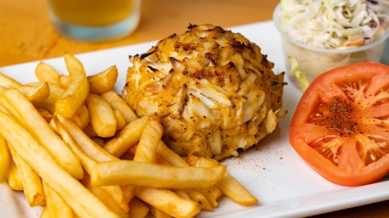 A crab cake on a plate with lettuce, tomato, and a cup of cole slaw