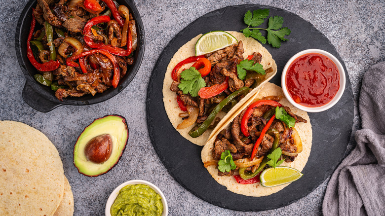 Tacos and fajita ingredients plated on black slabs.