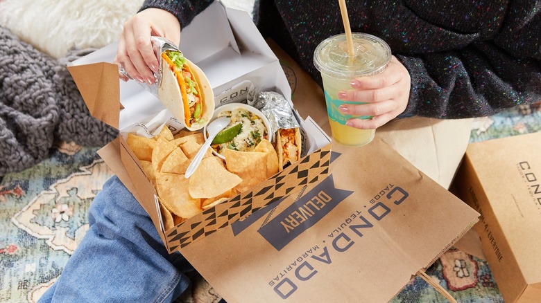Hands holding a Condado Tacos double-shell taco over a delivery box and a drink