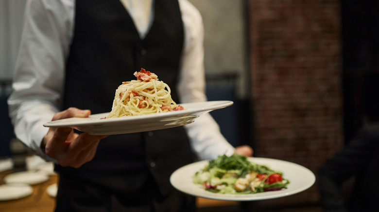 A vested waiter carries spaghetti and salad to a table.
