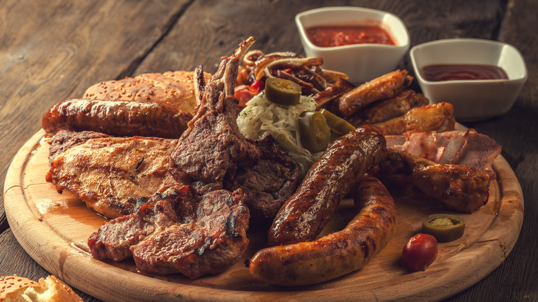 a barbecue meat plate featuring sausages, lamb, steaks, pork, chicken wings, and an array of sides, including cole slaw, dips, tomatoes, and jalapenos