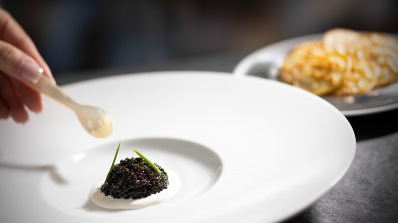 Someone uses a mother of pearl spoon to scoop up caviar on some creme fraiche, with blinis in the background