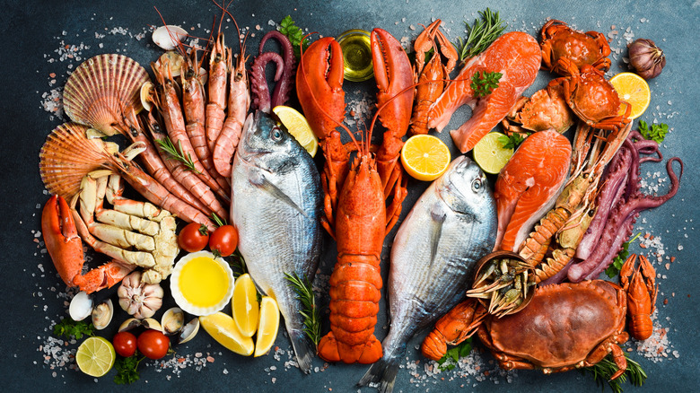 A selection of various seafood, from fish to lobster, as well as typical add-ins like lemon and garlic, are arranged artfully on a black table.