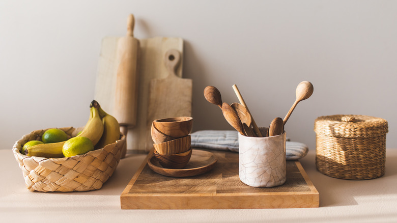 A set of wooden kitchen utensils sit on a wooden cutting board, next to a bowl of fruit in a warm, cream-colored kitchen