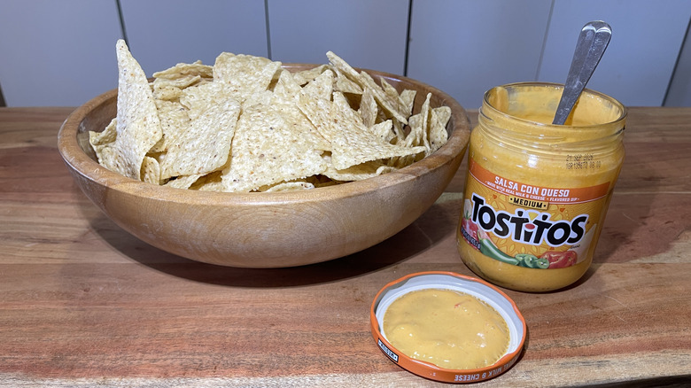 An open jar of Tostitos Salsa Con Queso with a spoon inside sits next to the lid and a bowl of tortilla chips.