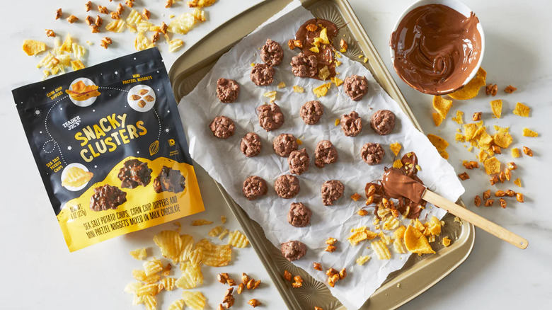 A bag of Trader Joe's Snacky Clusters sits next to a tray of clusters, crushed bits of salty snacks, and a bowl of chocolate