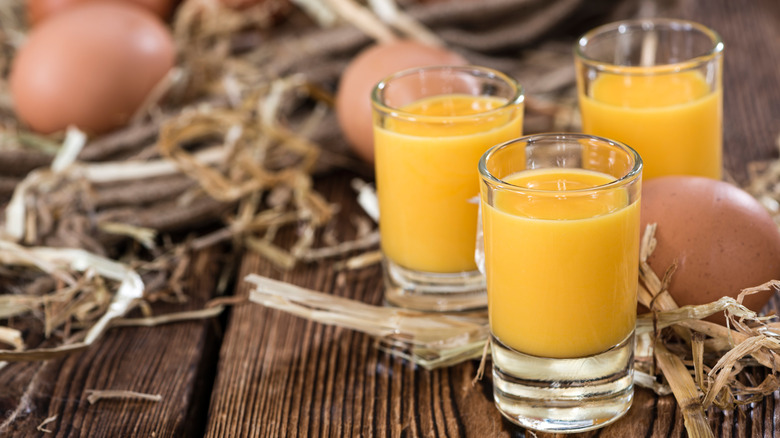 Three shots of Advocaat, a thick egg-based liqueur, sit on a wooden table with straw and eggs