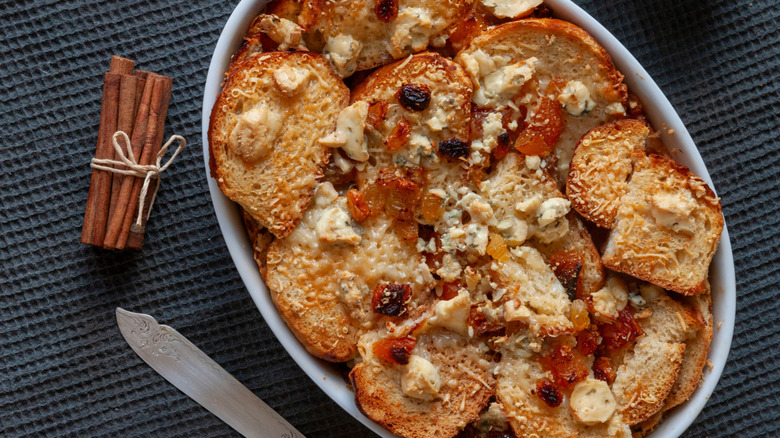 Mexican bread pudding with fruit and cheese rests in a white dish beside a knife and cinnamon sticks