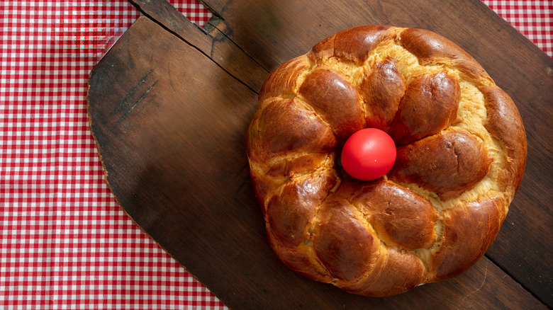 A round tsoureki, a Greek Easter bread decorated with a dyed red egg, sits on a wooden board