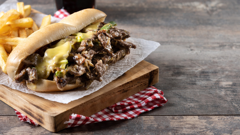 A Philly cheesesteak on a wooden platter with fries.