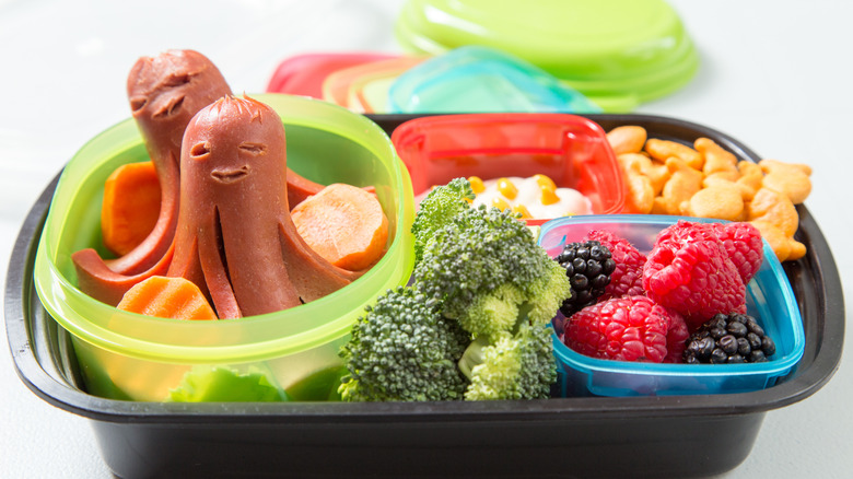 Cute little hot dog octopuses in a small container with sliced carrots, next to broccoli and fresh berries