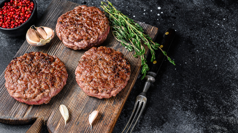 Cooked burger patties on a cutting board next to herbs and garlic