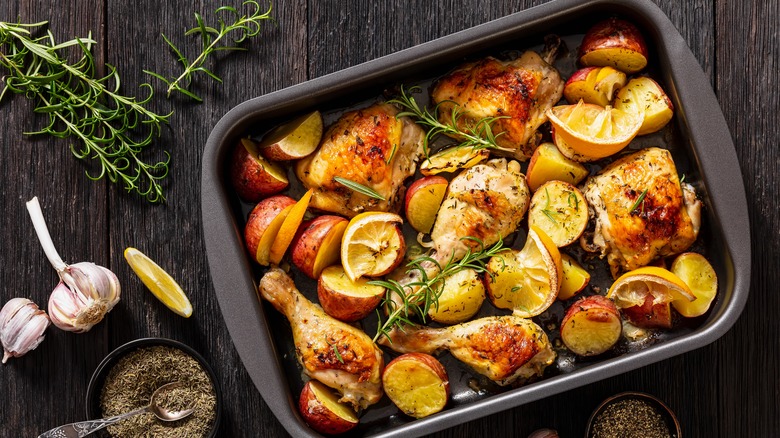 Sheet pan roasted chicken drumsticks and thighs with potatoes, rosemary, and caramelized lemons, surrounded by fresh rosemary sprigs, whole garlic, a lemon wheel, a bowl of dried thyme, and a bowl of cracked black pepper.