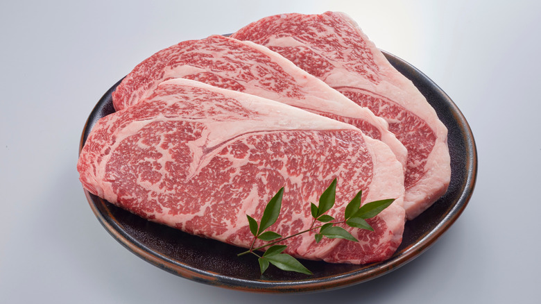 A plate of uncooked wagyu beef next to a sprig of herbs on a white background