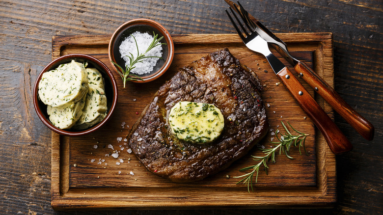 A well browned steak sits on a wooden cutting board with a pad of herbed butter next to a bowl of compound butter, salt, and fresh rosemary
