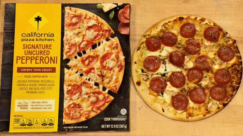California Pizza Kitchen Crispy Thin Crust Signature Uncured Pepperoni Pizza baked, with a slice cut out and pulled away from it, and placed on a wooden cutting board next to the box