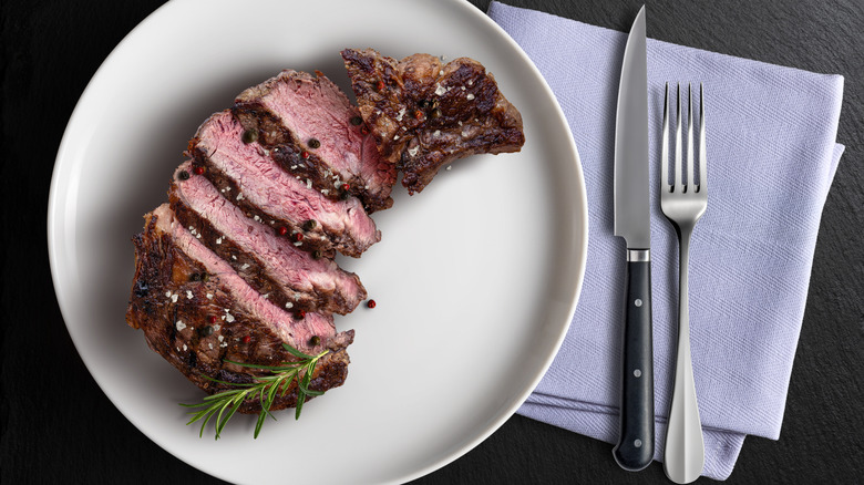 Sliced beef on a white plate garnished with a sprig of rosemary, with a napkin, knife, and fork to the side