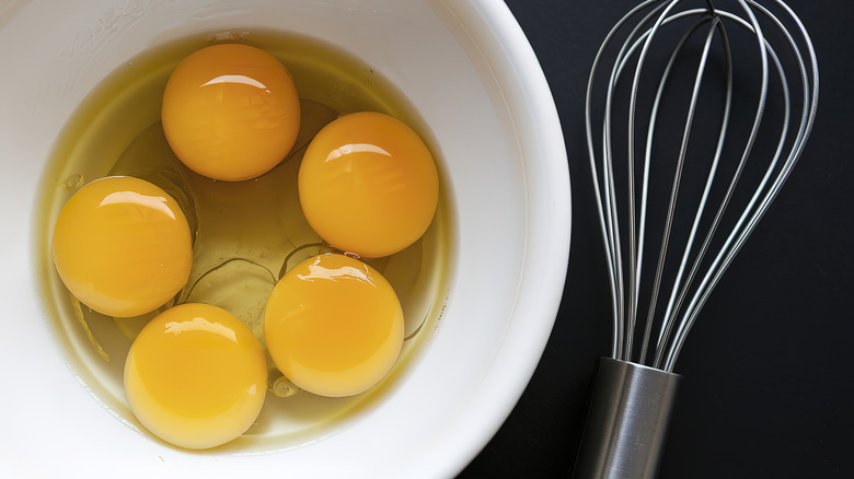 Five egg yolks sit in a white bowl with a whisk next to them
