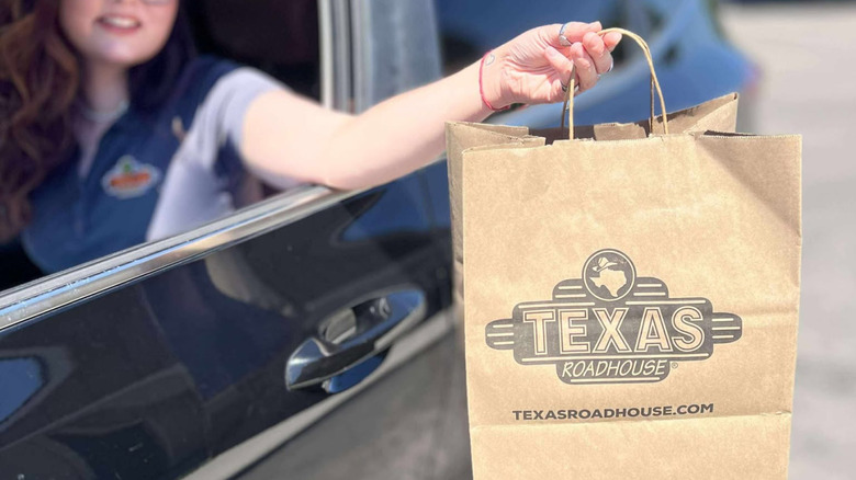 Someone in a car with their arm extended out a car window holds a Texas Roadhouse takeout bag.