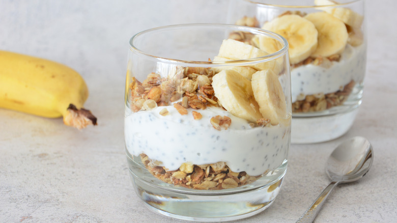 Granola, yogurt with chia seeds, and banana layered in two glasses alongside a spoon and a banana.