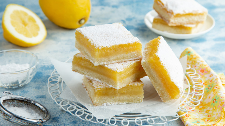 Focus on lemon squares stacked on a plate with powdered sugar, lemons, additional lemon squares and a floral napkin surrounding. All on a blue and white table.