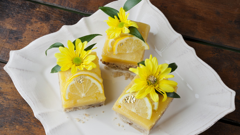 Top view of three lemon squares decorated with lemon slices and yellow flowers on top. All on a white square plate, and a wooden table.