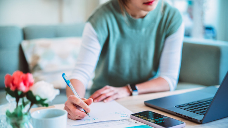 A woman sits at a computer in front of a laptop, writing in a notebook