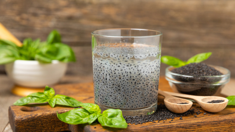 Soaked basils seeds in a glass of water next to fresh basil leaves and spoonfuls of spilled basil seeds