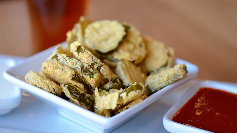 Golden brown fried pickles in a bowl next to two dipping sauces.