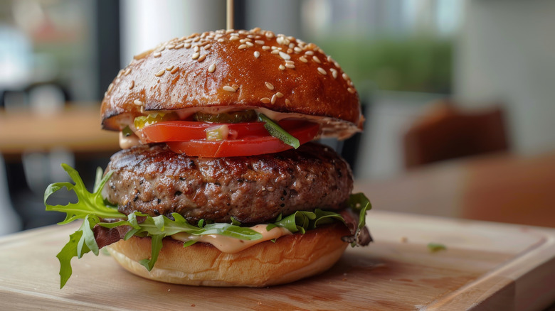 A burger with a rustic sesame seed bun, pickles, tomato, arugula, and burger sauce sits on a cutting board