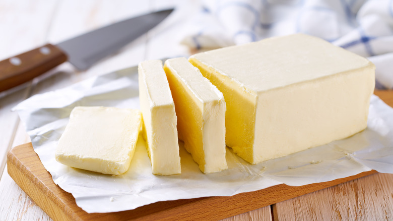 An open package of butter sliced on a cutting board, a knife behind it.
