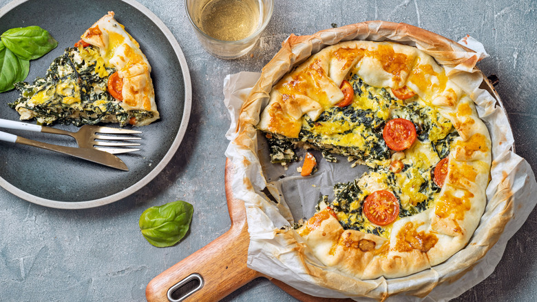 A savory spinach and tomato quiche has a slice taken out, which sits on a plate with a fork and knife as well as some loose basil leaves