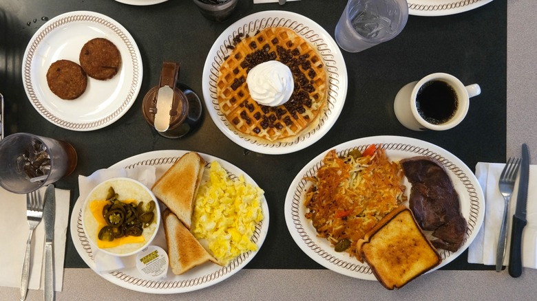 A feast laid out including waffles, sausage, eggs, toast, grits, and a plate with steak, hasbrowns, and toast.