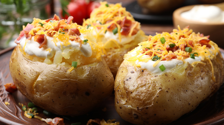 Three baked potatoes on a plate, topped with sour cream, bacon, scallions, and cheddar cheese