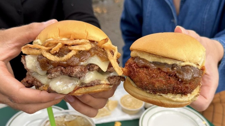 Two people holding Shake Shack burgers, one with caramelized mushrooms and onion strings, another with a fried patty.