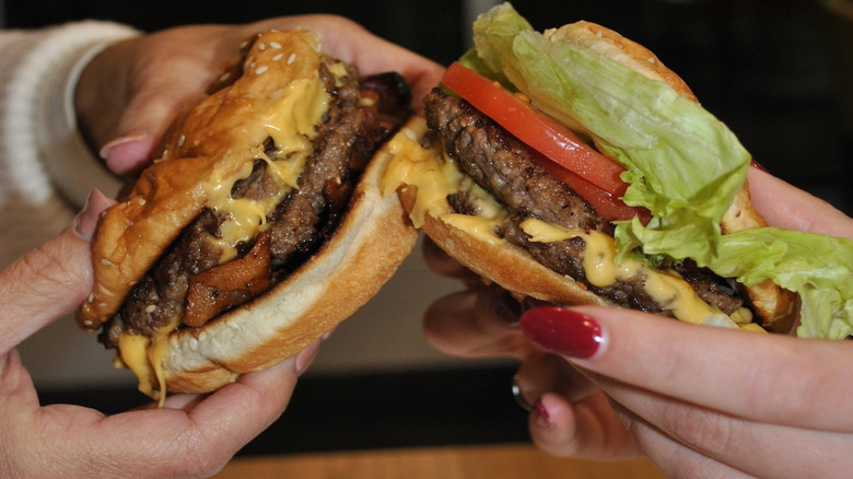 Five Guys bacon cheeseburger and cheeseburger with lettuce and tomato held by two pairs of hands.